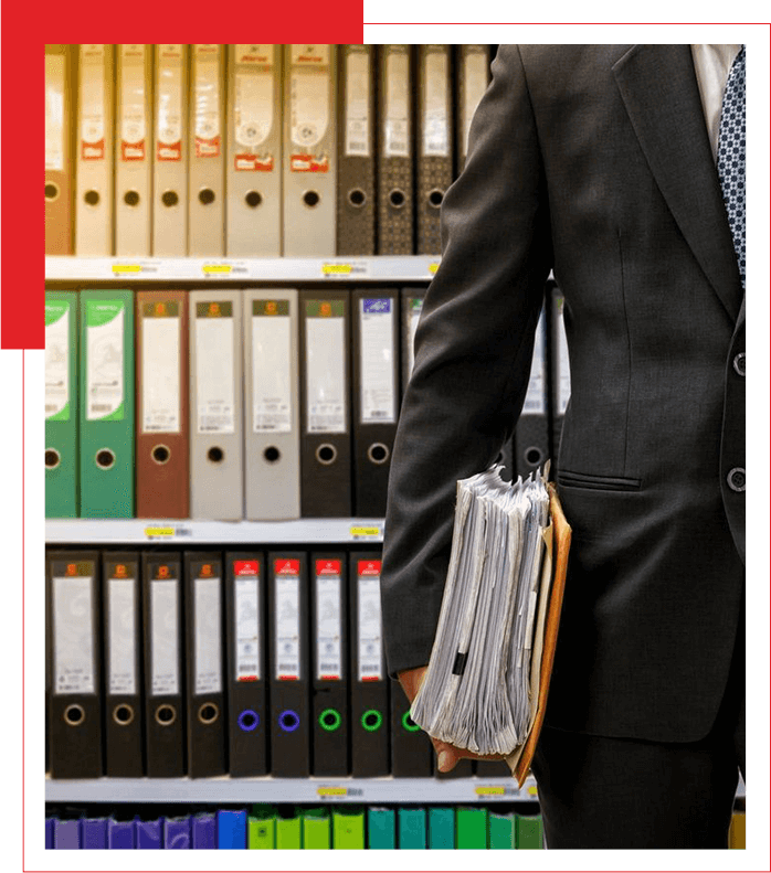 A professional in a suit holding a stack of documents with a shelf of binders in the background.